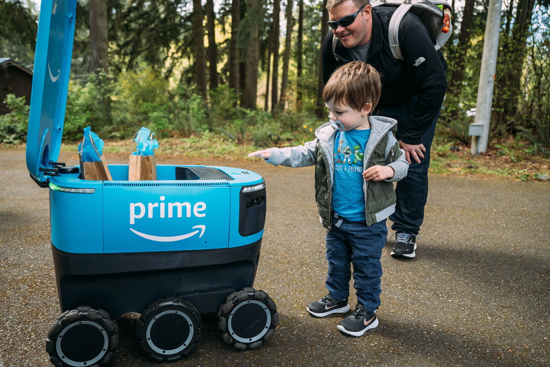 A young boy reaches toward a Scout delivery robot. His father smiles as he leans toward him. 