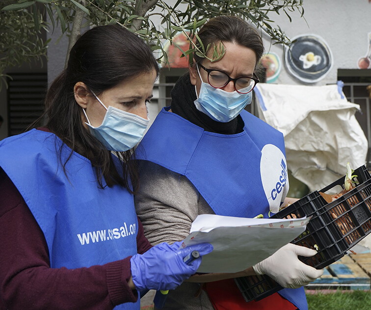 Con el proyecto Stop Coronavirus la ONG Cesal atiende a más de 400 familias en Madrid. Dos voluntarias con el chaleco de Cesal, guantes y mascarilla están mirando el papel que una de ellas sujeta con la mano. La otra sujeta una caja de plásitco de color negro que tiene cebollas. Las dos tienen el pelo largo y la que sujeta la caja de cebollas lleva gafas. Están en el exterior. 