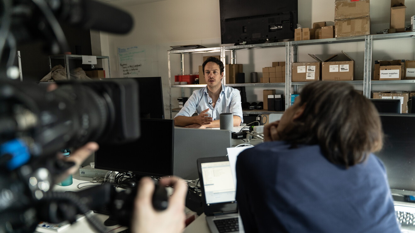 Francesco Piai, Beta Program Manager. Francesco está en su lugar de trabajo con el equipo de rodaje. Él está en el fondo, sentado en su mesa y con un dispositivo Echo. Rodeado de cajas, enfrente tiene a un cámara y a una periodista.