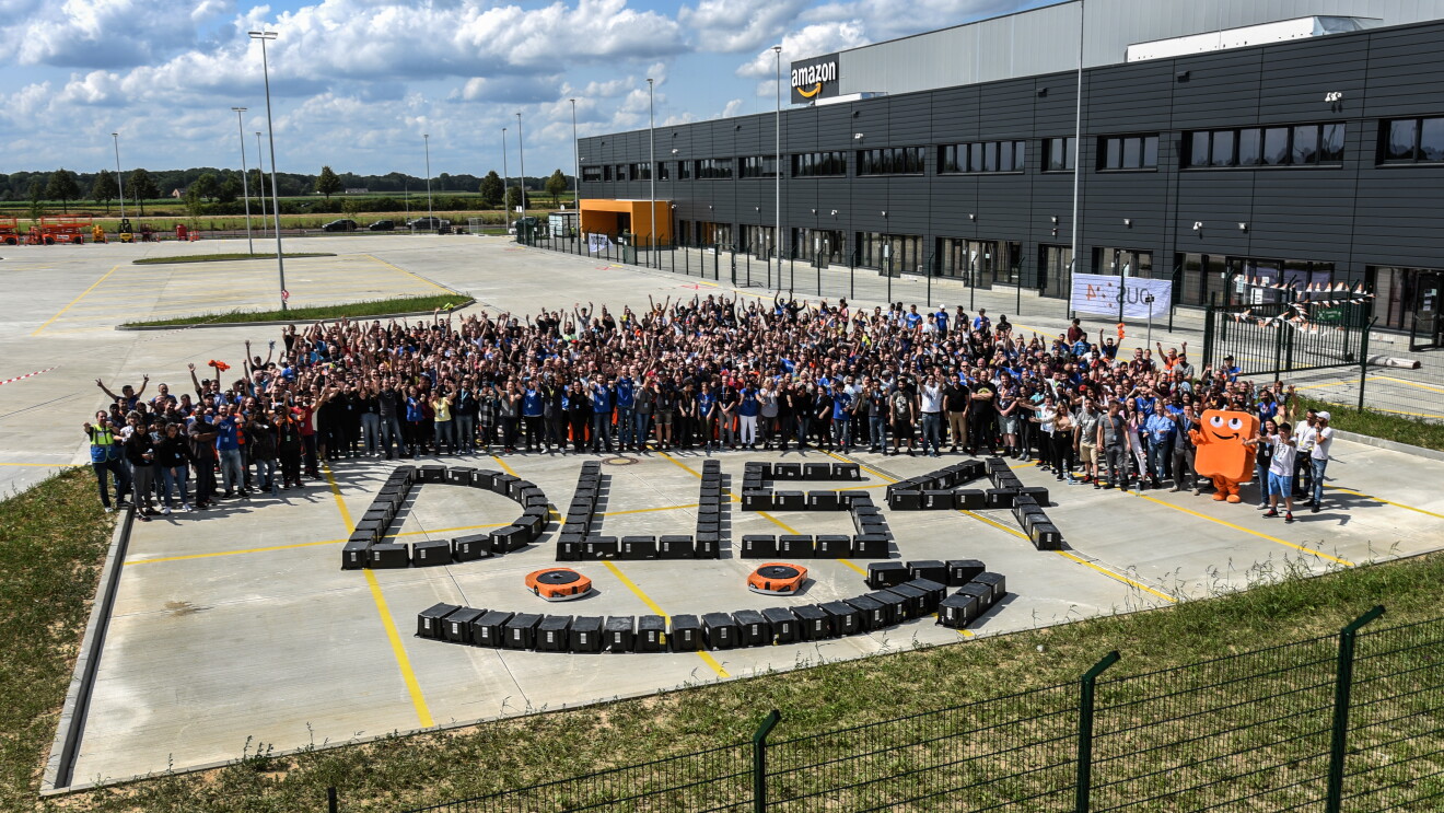 Ein Gruppenbild mit Mitarbeitern vor dem Amazon Gebäude. Vor ihnen wurde aus schwarzen Transportkisten der Schriftzug DUS4 geformt, zusammen mit dem Amazon Logo Pfeil und 2 orangenen Transportrobotern.