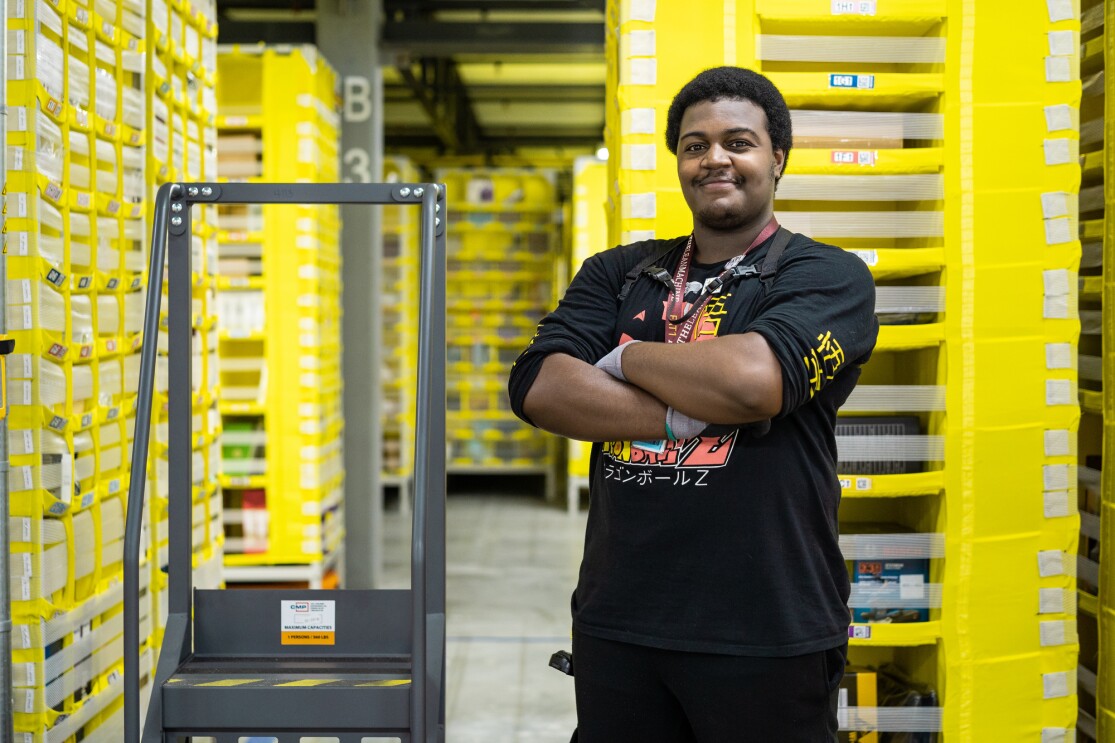 An Amazon employee, wearing a long-sleeve tee shirt, black pants and a lanyard stands with his arms crossed. Next to him is a step ladder. Behind him are tall yellow storage stacks. 