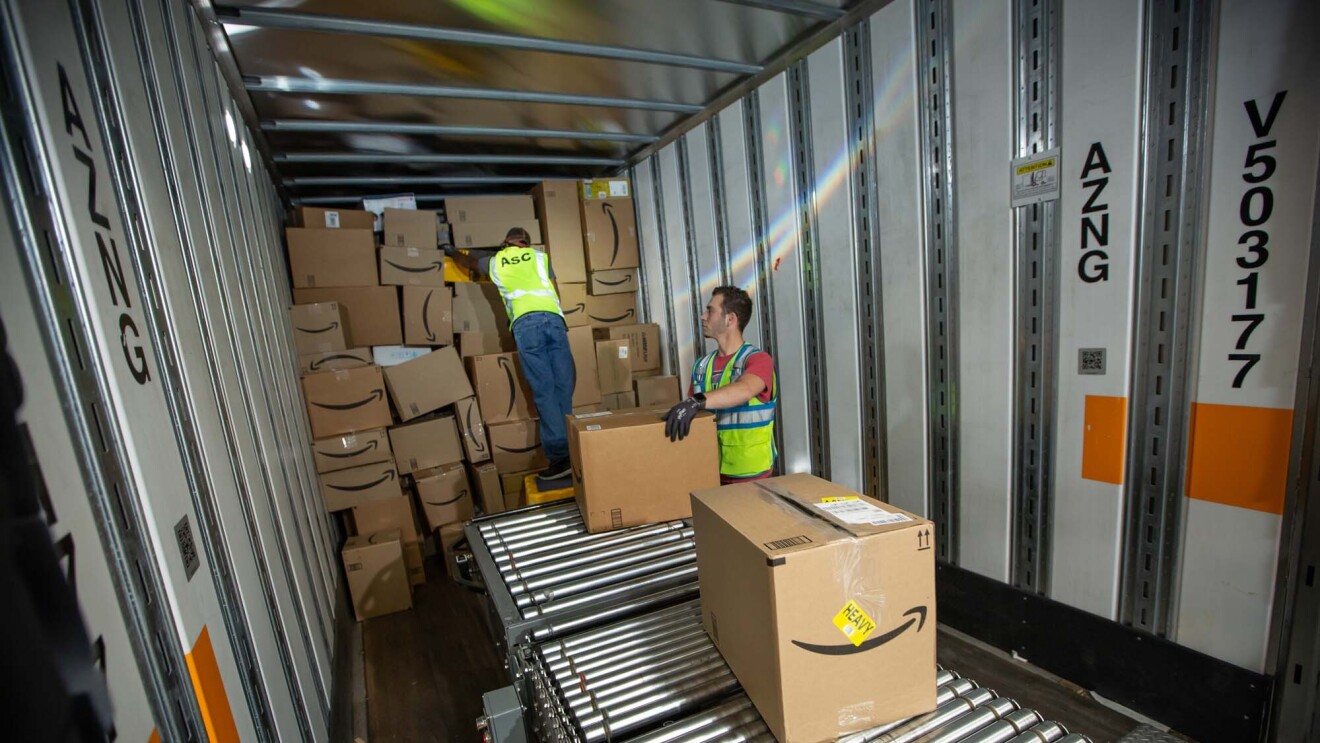 Two Amazon associates receive packages from a conveyor belt, as they prepare to place them on a trailer for distribution at a Columbus, Ohio fulfillment center, CMH2