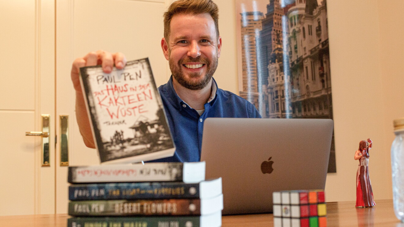 Paul Pen mostrando la traducción al alemán de su novela La casa entre los cactus. En su habitación de trabajo Paul sujeta su novela traducida al alemán con su mano derecha. Delante tiene el ordenador, sus otras novelas traducidas. De fondo hay una puerta y un poster colgado en la pared. Paul está mirando sonriendo a cámara con una camisa azul. 