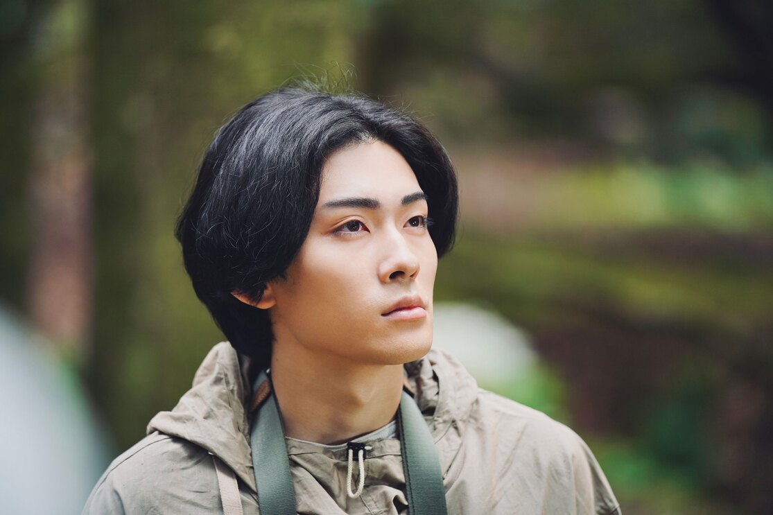 薄いカーキ色のジャケットを着た人物が少し上を見つめているクローズアップ。緑のストラップを首にかけている。背景はぼやけた緑の自然。