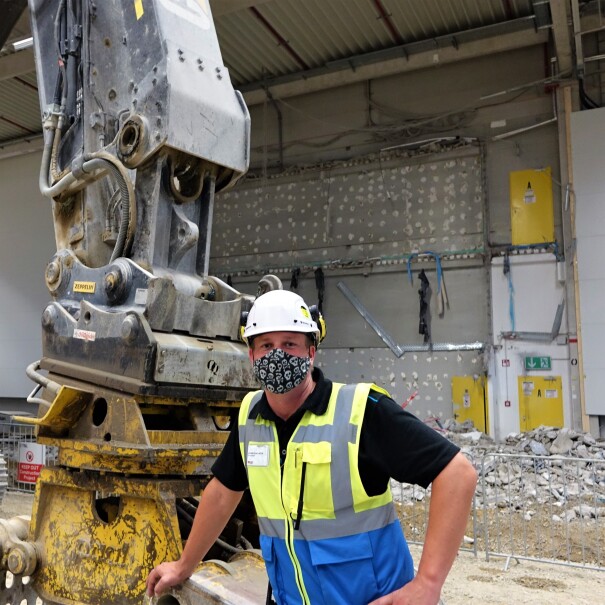 Oliver Reisenhofer mit Maske und Helm neben einem Bagger auf der Baustelle