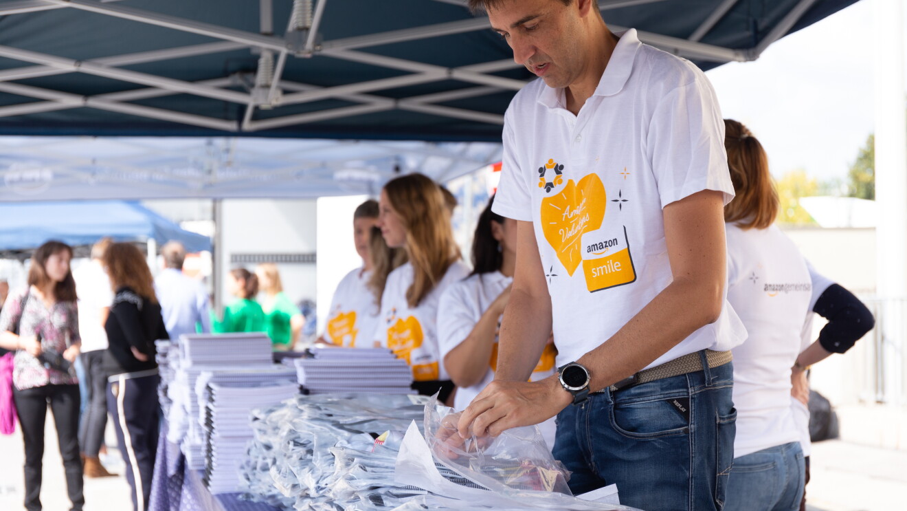 Amazon Mitarbeiter helfen beim Verteilen von Schulmaterial, wie Bücher, Hefte, Mappen. Alle tragen das AmazonSmile T-Shirt mit einem orangenen Herz auf der Vorderseite.