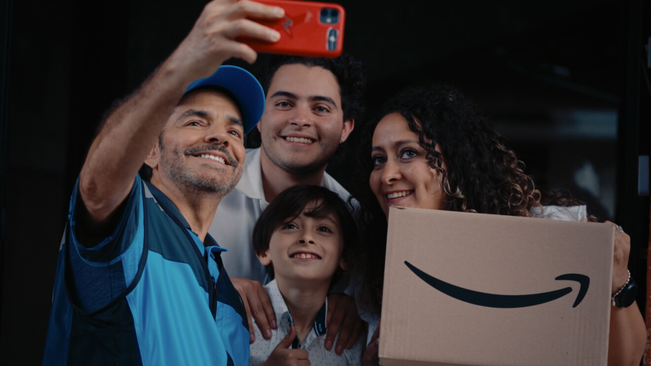 El actor Eugenio Derbez caracterizado de repartido realizando una entrega a una familia. En la imagen el actor se está sacando una foto con una familia de unos padres y un hijo. La madre agarra una caja de Amazon.