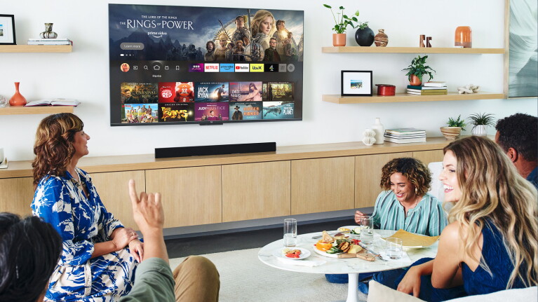 A group of people gathered in a living room with a Fire TV mounted on the wall and food on the table.
