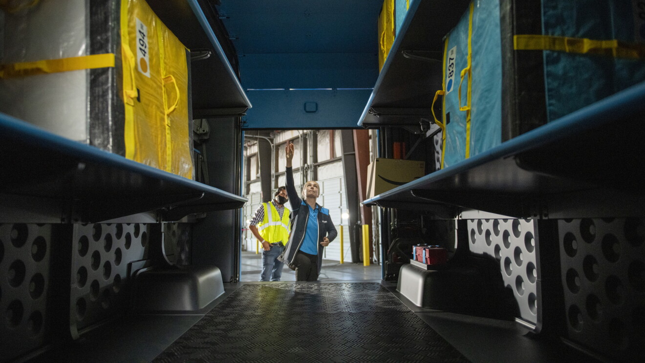 Two people stand at the back of a delivery vehicle and look at the shelving designed to hold Amazon customer orders.