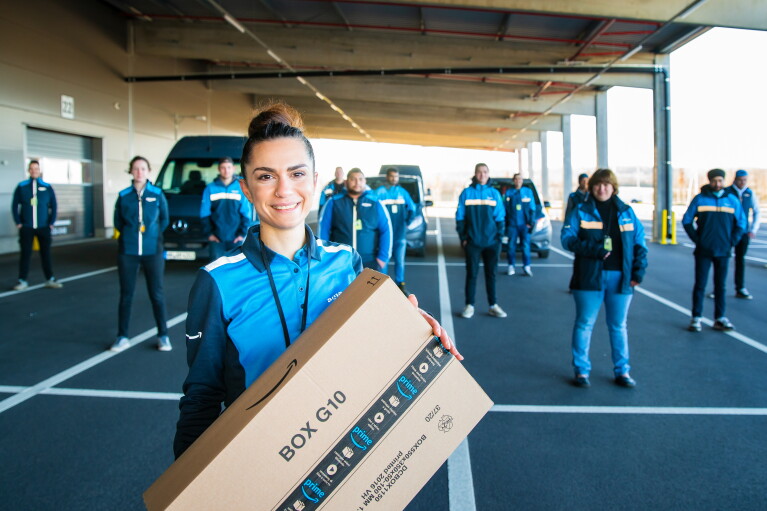 Eine zierliche junge Frau steht im Vordergrund mit einem Amazon Paket, hinter ihr sind 30 Mitarbeiter:innen verteilt auf einem Parkplatz zu sehen. Alle tragen Arbeitskleidung im Amazon Fahrer:innen Branding.