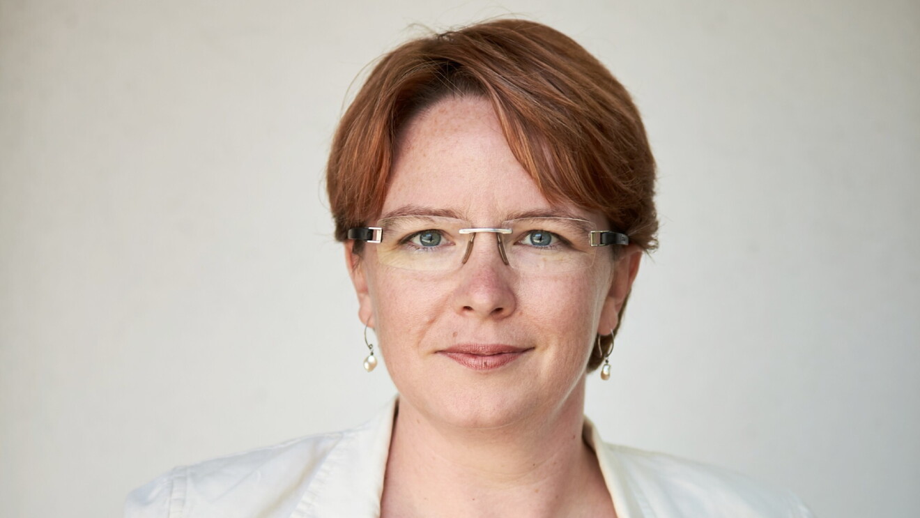 A woman smiles for a headshot photo in front of a white background while wearing a white blazer and glasses.