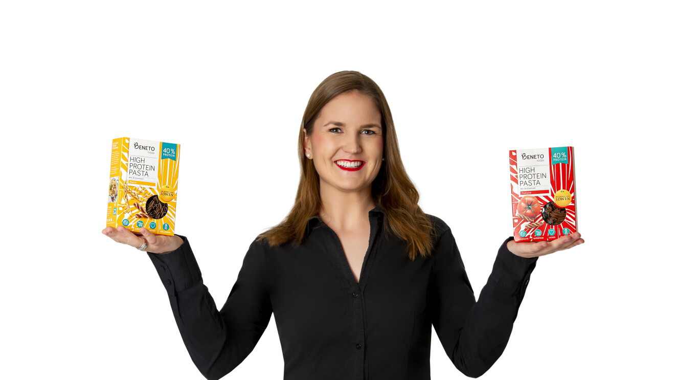 Eine Frau mit schwarzer Bluse lächelt in die Kamera. In ihren Händen hält sie Nudelpackungen. 