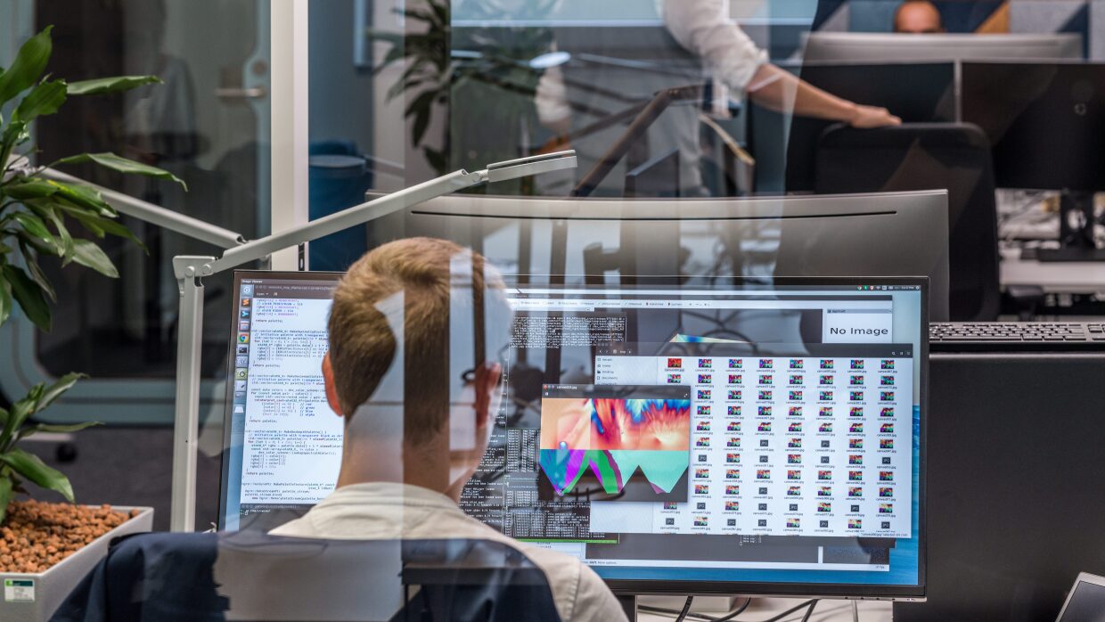 Amazon Research Center en Tübingen, Alemania. Septiembre de 2019. En una oficina de Amazon sen tres meses correlativas y en el fondo una persona cara a cámara trabajando en un ordenador con la pantalla que le tapa casi toda la cabeza. En segundo plano un hombre con una camisa y una armilla, con una mano apoyada en una silla y de espaldas al objetivo. En primer plano aparece un hombre de espaldas delante de una gran pantalla de ordenador. 