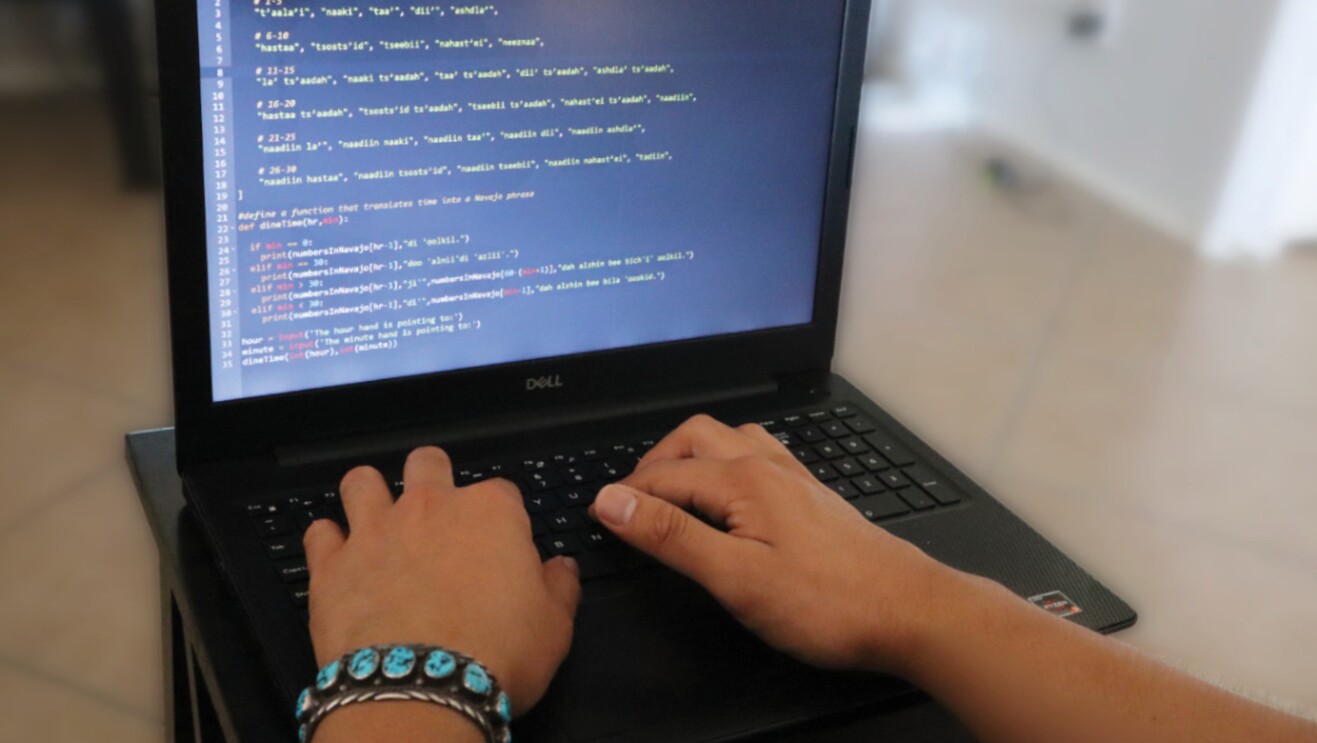 An image of a laptop with a code shown on the screen and two hands typing on the keypad.