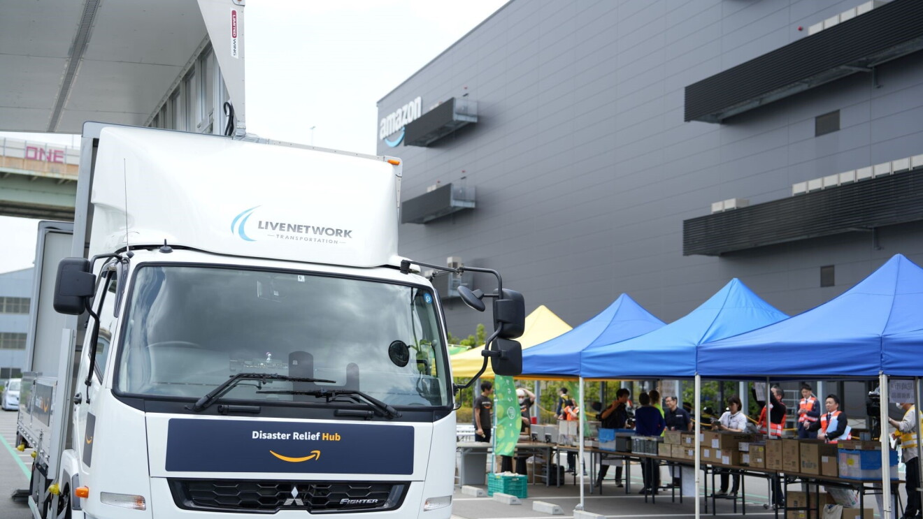 An Amazon Disaster Relief Hub vehicle stops in front four tents where several people are organizing supplies and relief items