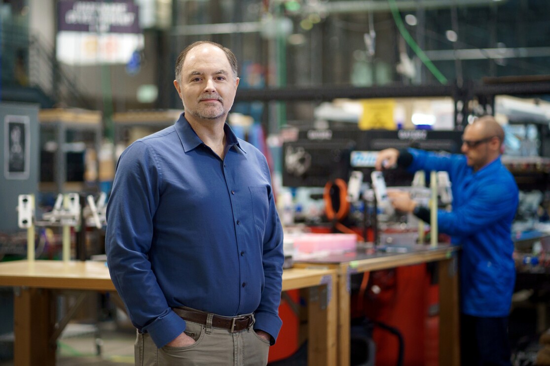 A man wearing a blue button down and jeans stands with his hands in his pockets in robotics workspace.