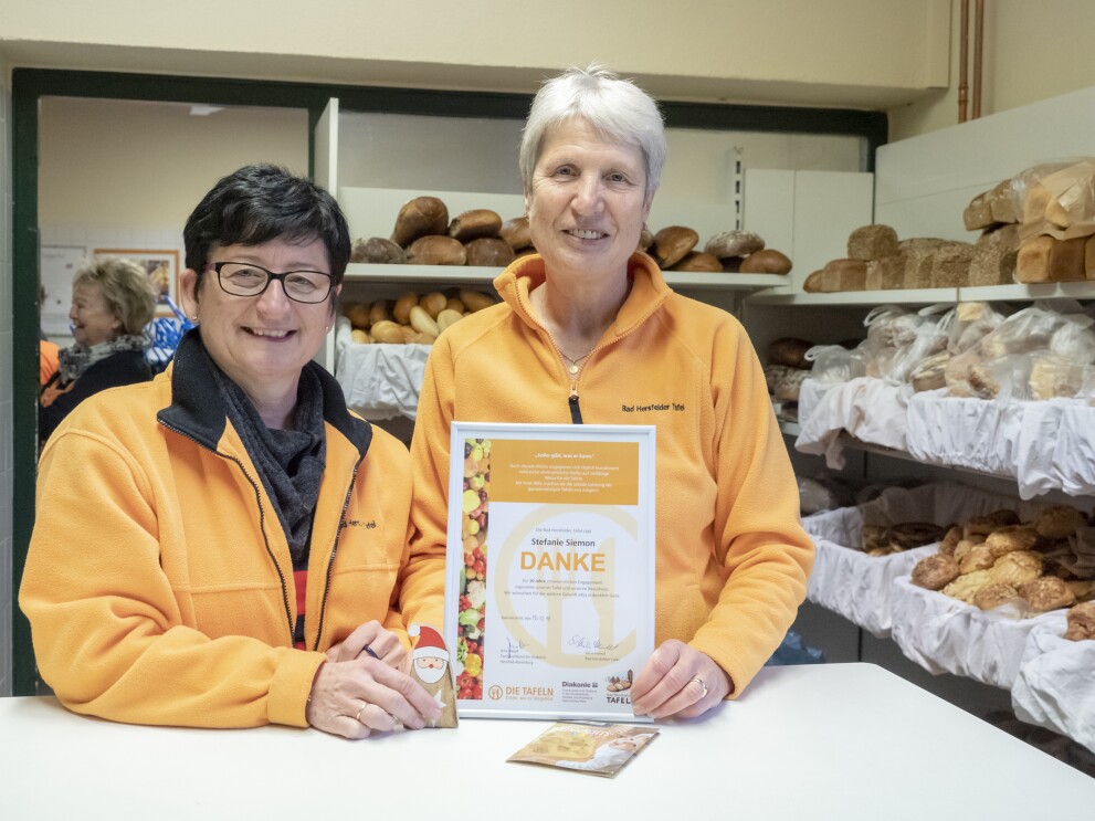Zwei Frauen, beide mit orange-farbigen Fleece-Jacken mit dem Aufdruck "Bad Hersfelder Tafel". Die rechte trägt einen dunklen Kurzhaarschnitt und eine Brille mit dunklem Rahmen. Neben ihr eine ältere grauhaarige Dame. vor ihnen steht ein Bilderrahmen auf dem Tresen mit einer Dankesurkunde.