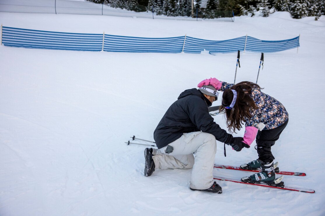A father helps his young daughter click her ski boots into her skis.