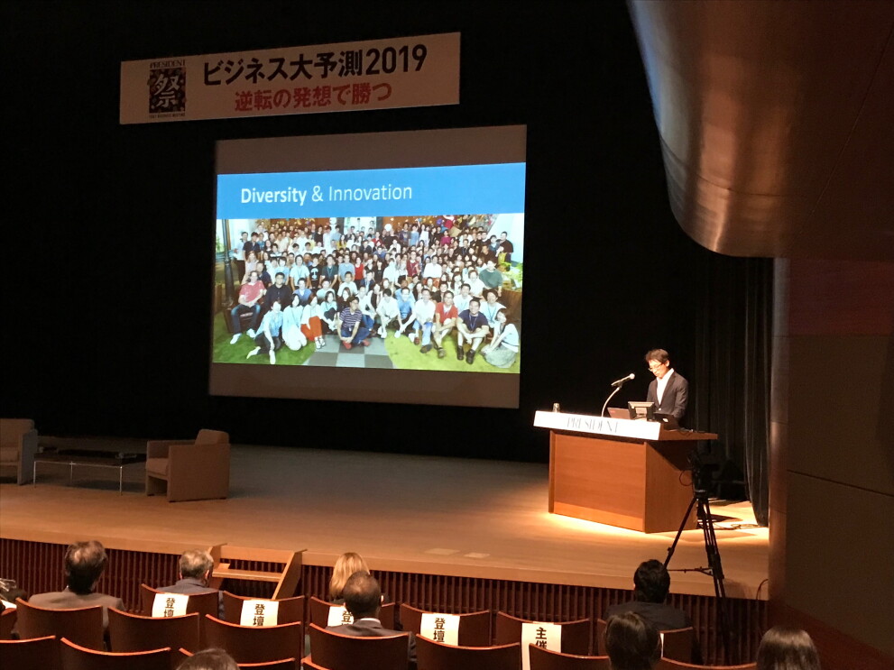 薄暗いホールのステージ上で話をする登壇者。ステージ奥のスクリーンに集合写真が投影されている。ステージ下に登壇者用の椅子が並ぶ。