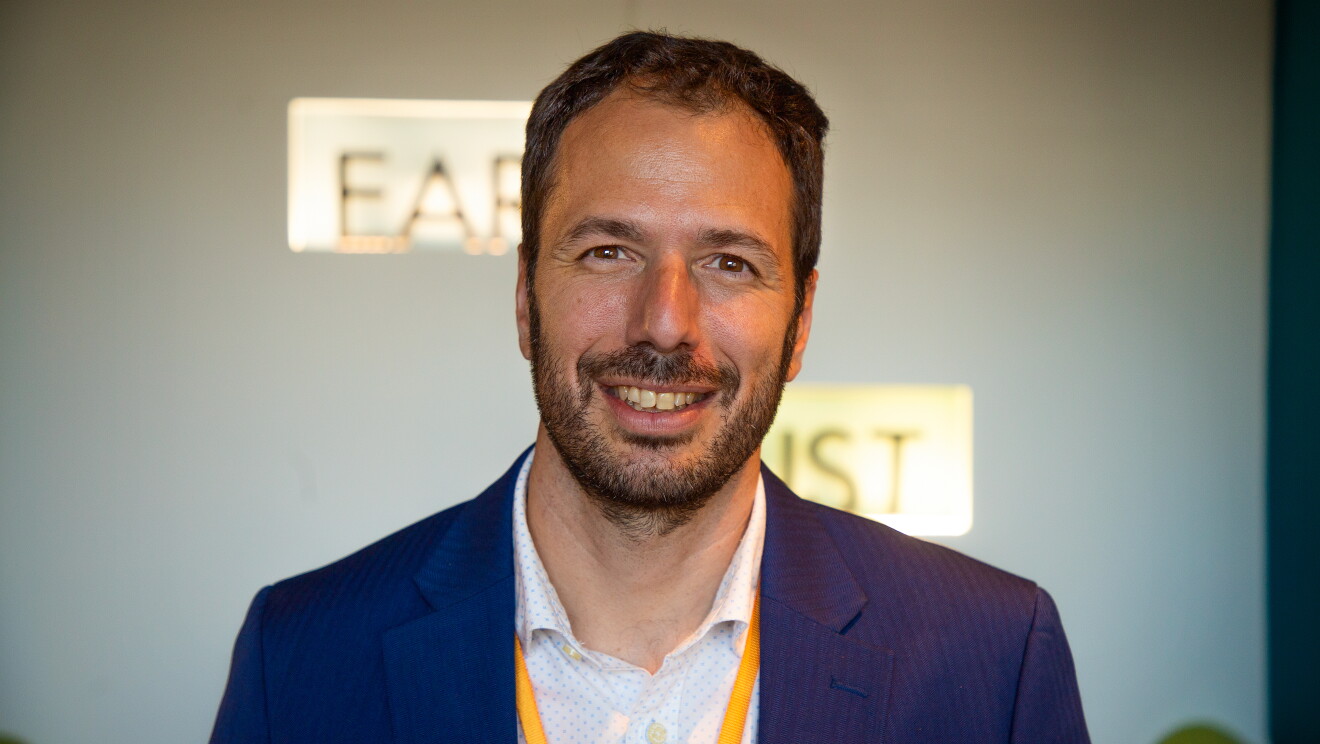 Juan Barbero_Program Manager de Despega. Aparece Juan en un plano corto por los hombros. El pelo negro corto con barba negra de cuatro días. Americana azul, camisa blanca con motivos de color azul y collar del badge de color amarillo. De fondo pared blanca y una letras iluminadas que no se pueden leer porque están difuminadas. 