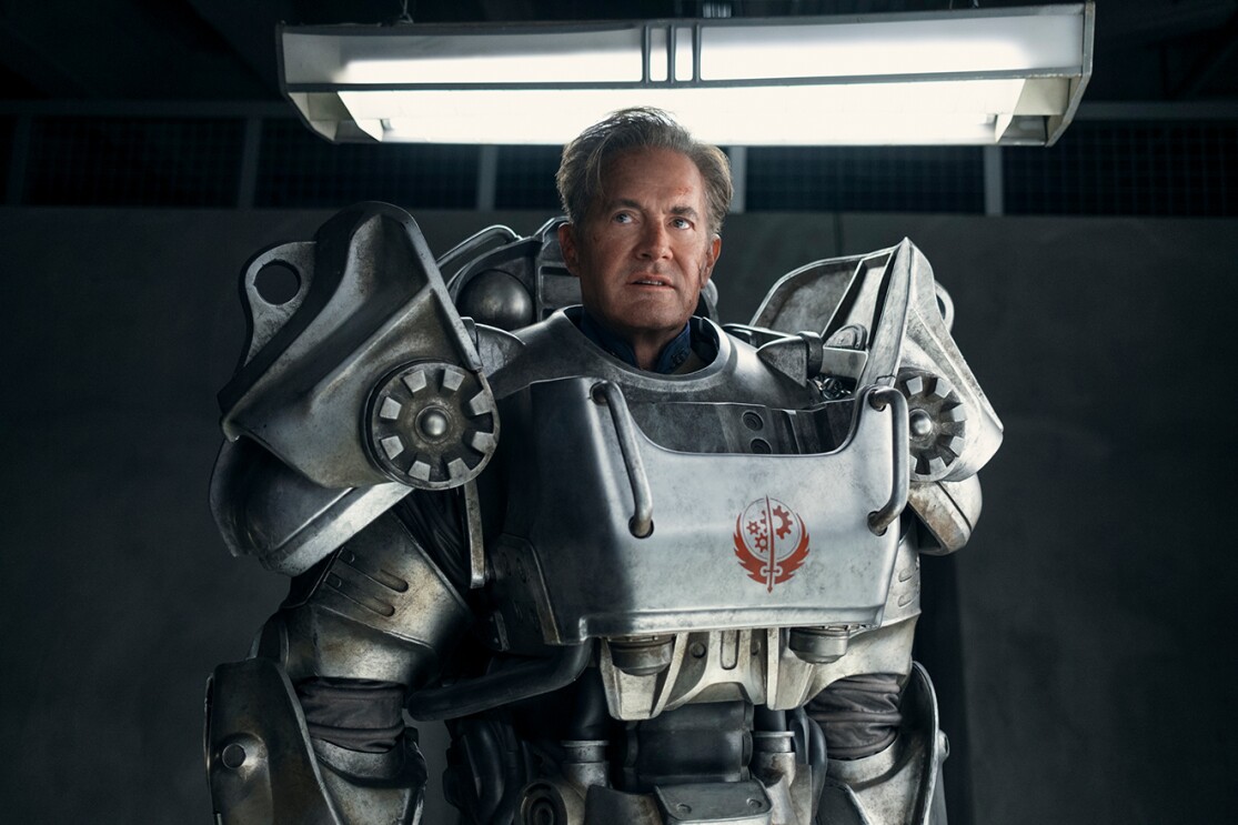 薄暗い屋内に立つ、鈍い銀色のパワーアーマーを着用した中年男性。肩当てパーツが大きく、胸部プレートに赤い紋章が書かれている。頭上に白く光る蛍光灯。