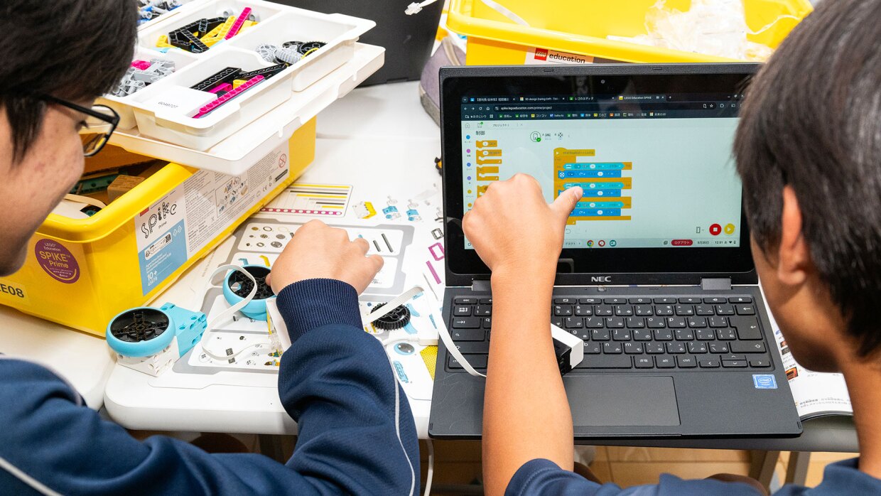教室で、2人の生徒が机に向かって作業している。白い机の上には黄色いボックスと白いトレー。右の生徒はノートパソコンを使ってプログラミングを行い、左の生徒が水色と白のパーツで組み立てた構造の動きを確認している。パーツの下に設計図のような紙。