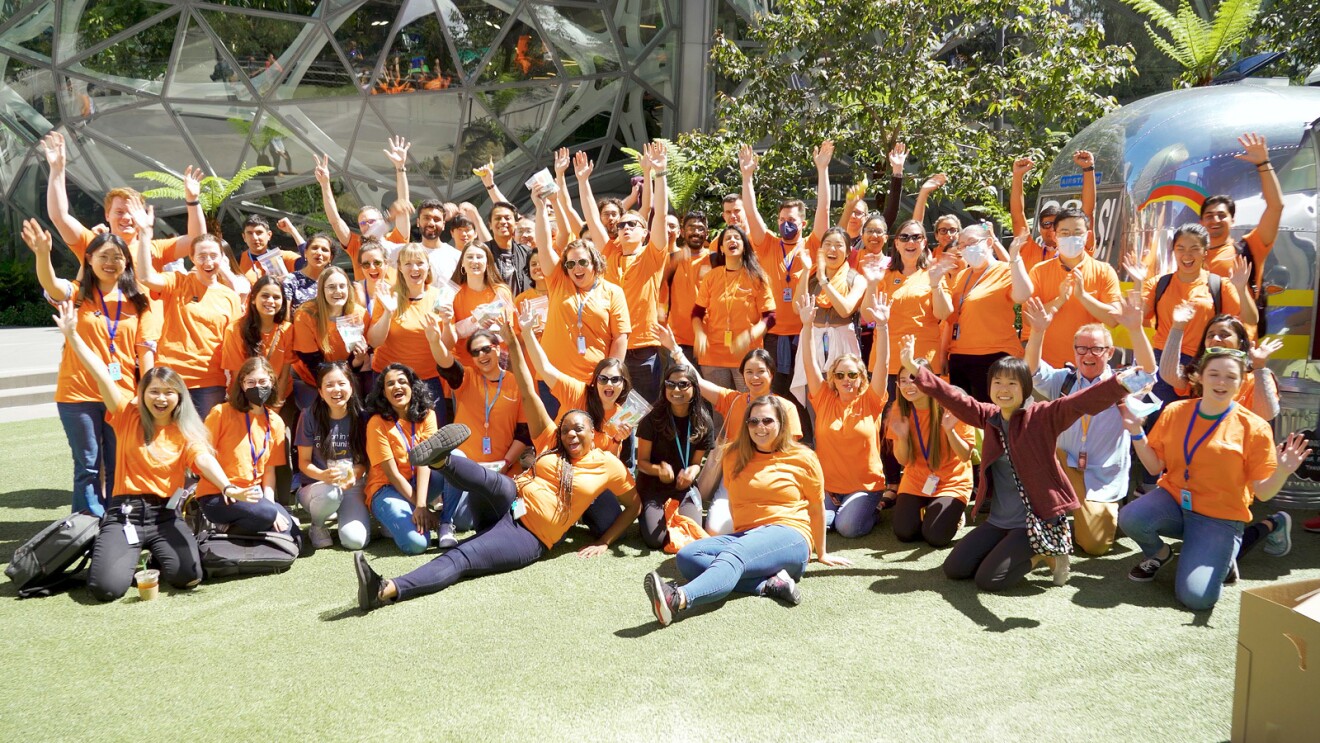 A big group of Amazon employees wear orange T-shirts and raise their hands in celebration as they pose for a picture outside of Amazon's headquarters.