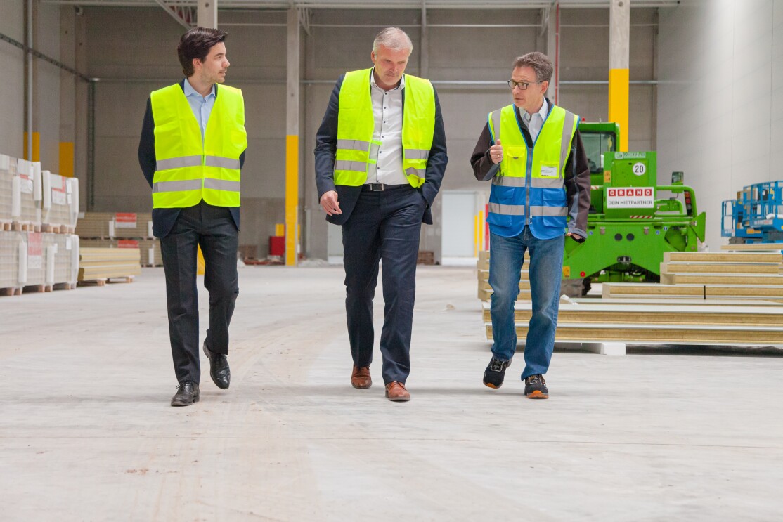 3 Männer in gelben Sicherheitswesten gehen Seite an Seite durch eine Halle.