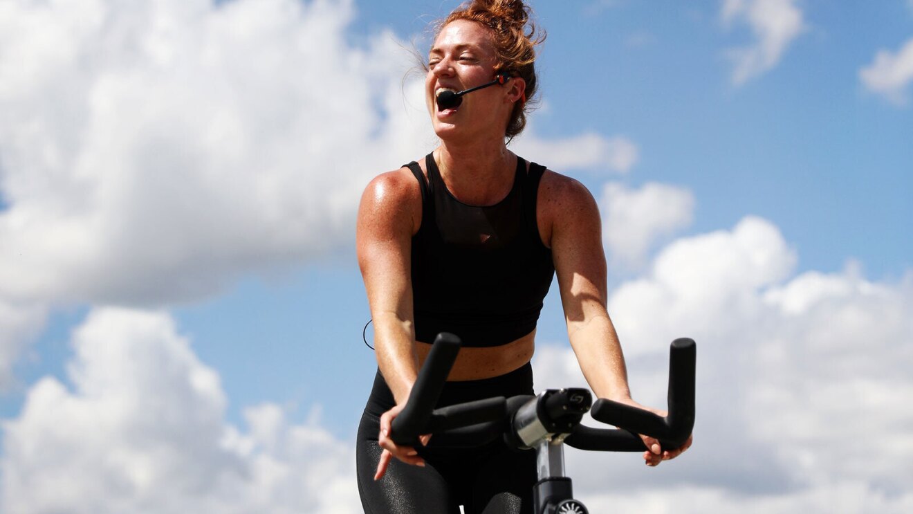 An image of a woman teaching a spin class outside. She is riding a stationary bike wearing a microphone and smiling while saying something to the class. There is a blue sky with light, white clouds behind her in the distance.