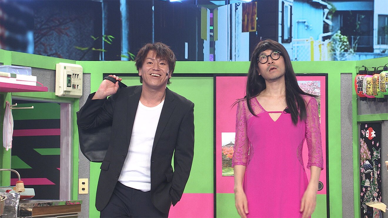 ふすまや壁の一部が明るい黄緑とピンクに塗られたコント用のセットに、2人の男性が立っている。左側は黒いジャケットで鞄を肩に背負い、右側はピンクのワンピースと長髪で女装している。