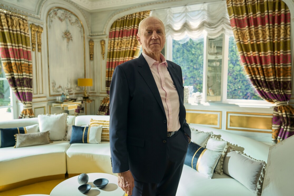 Elderly man in suit standing in opulent room with ornate curtains