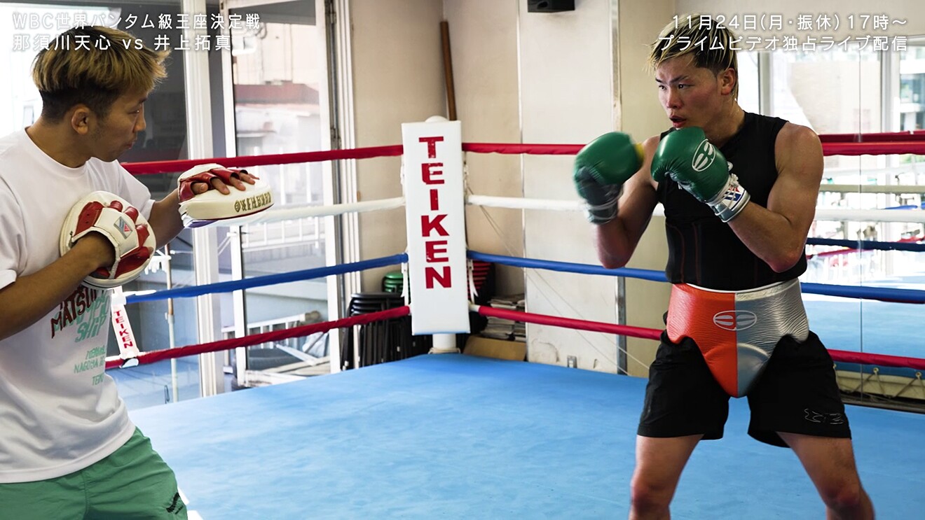 ジムの室内、青いマットのボクシングリングで向かい合う2人の男性。左側はトレーナーでミットを着用、右側はグローブと股間ガードを着用した選手。コーナーポストに「TEIKEN」の文字。