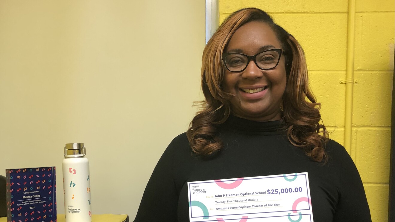 A woman smiles for a photo while holding two checks she earned as an award a recipient of Amazon Future Engineer's Teach of the Year award. One check is for $25,000 and the other is for $5,000.