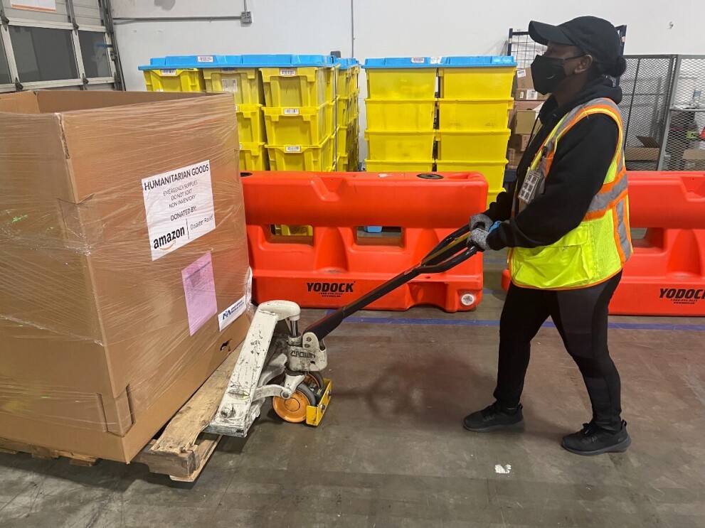 A woman wearing a yellow safety jacket and a face mask pushes a box that reads 'humantarian goods' with a pallet jack.