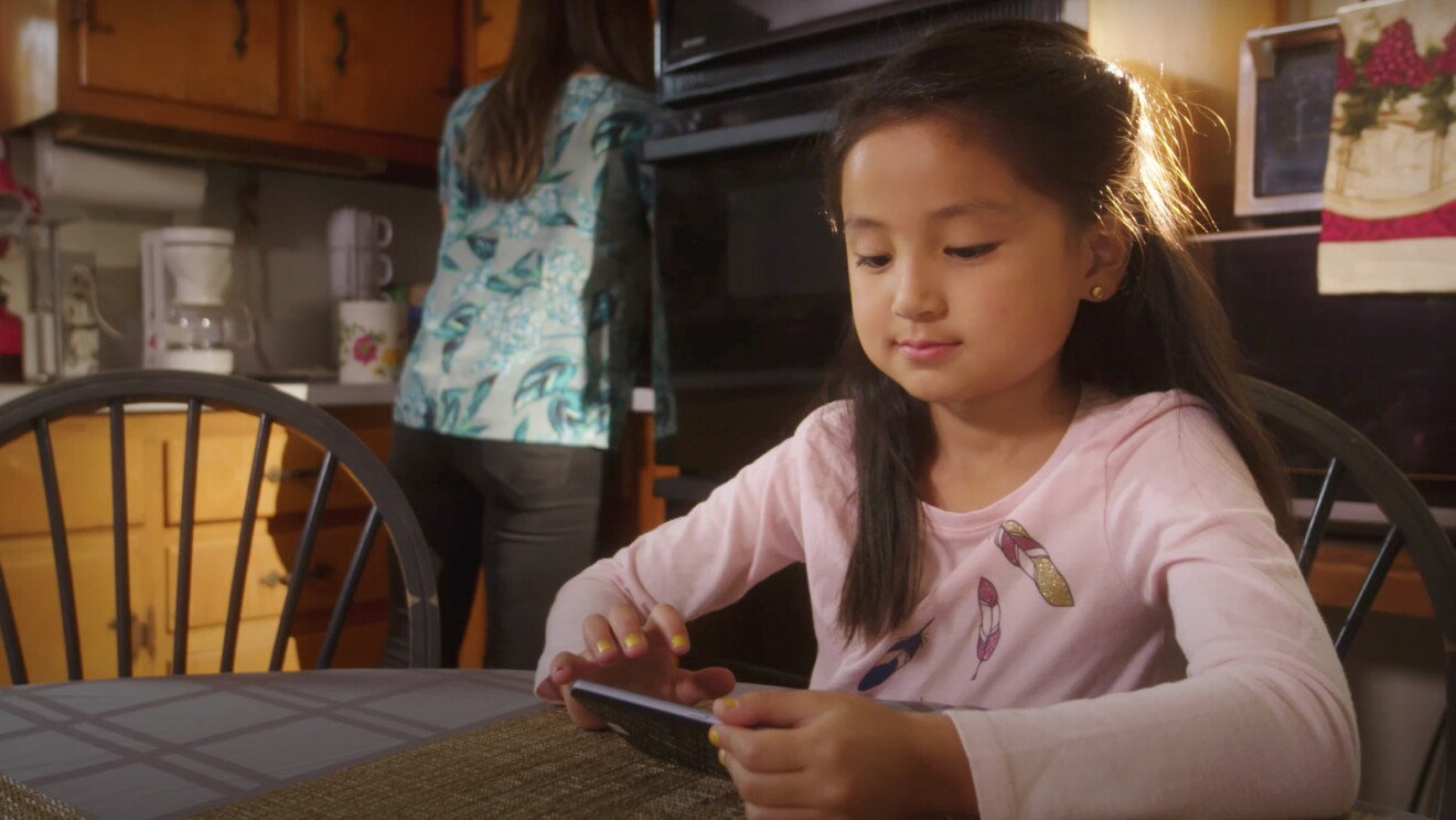 A photo of a child using an app on a mobile device while sitting in a kitchen at a table.