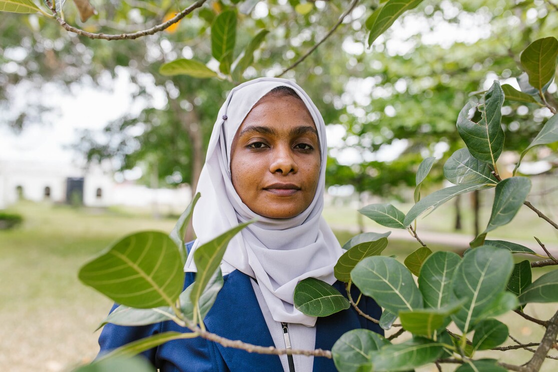 A young woman looks at the camera, from behind leaves on a tree branch