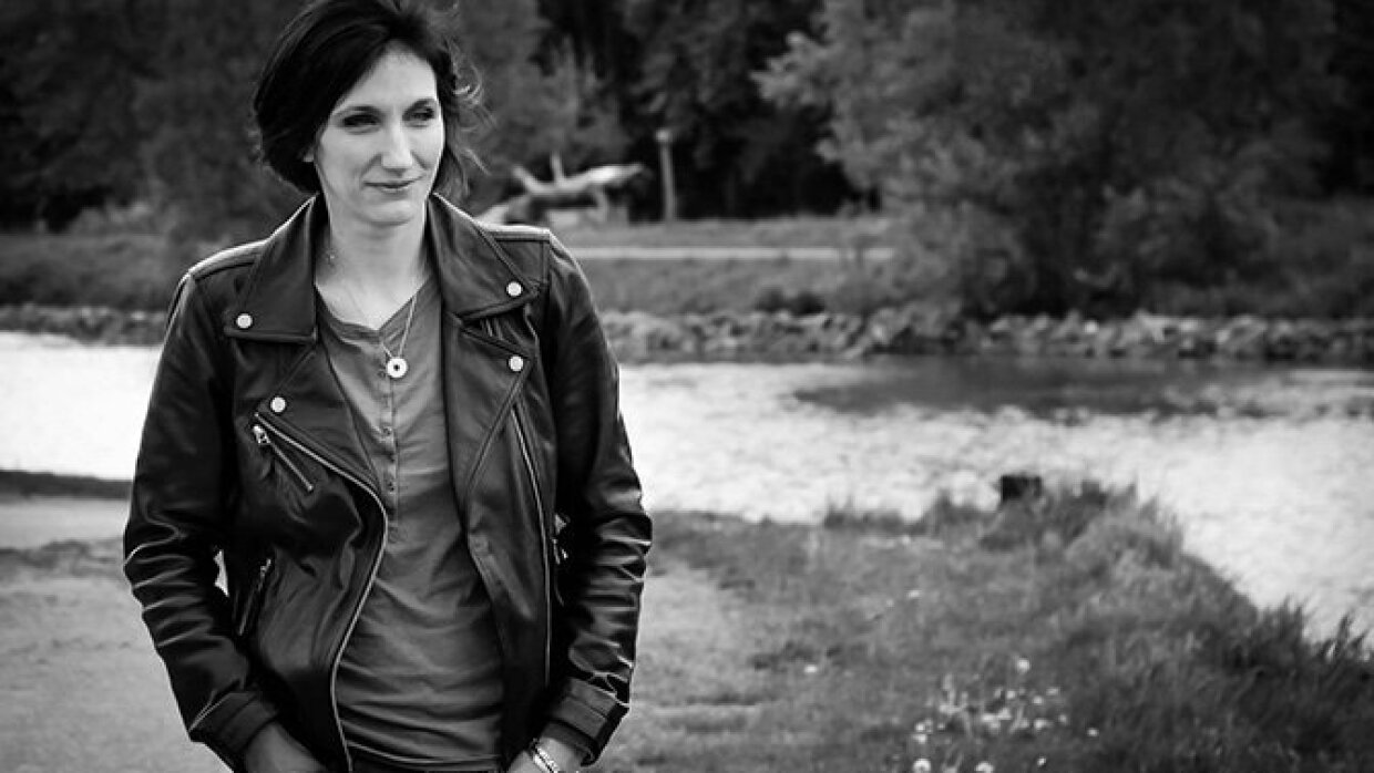 Amélie Antoine se promène dans la campagne en blouson de cuir, derrière elle se trouvent une rivière et des arbres. C'est une photo noir et blanc.