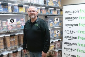 An image of a person standing in front of an Amazon Fresh banner with packages of food in the background. 