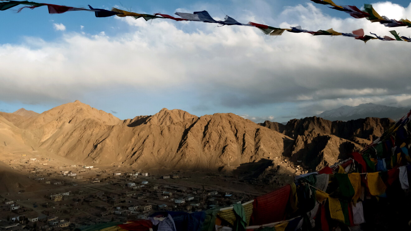 Prayer flags flutter in a breeze. In the background there is a valley with buildings. Mountains rise up in the distance.