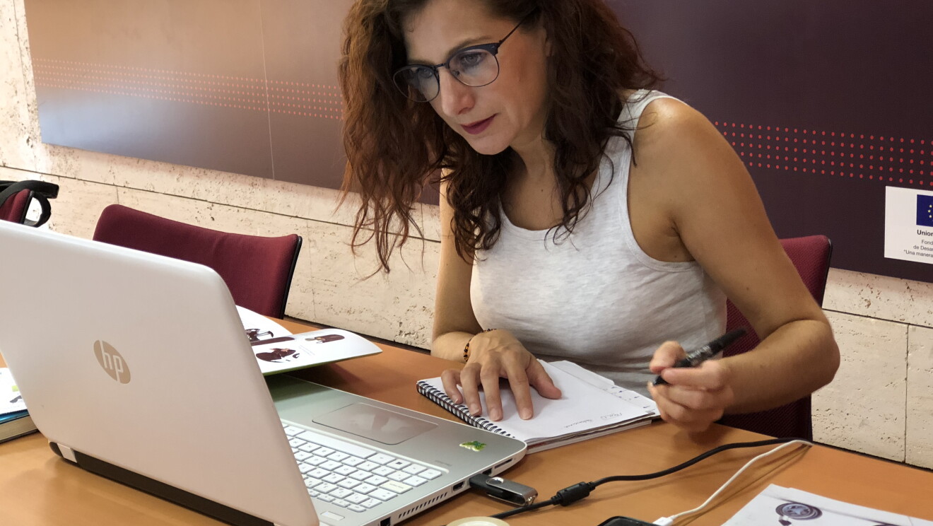Irene, creado de Andone Dynamic Walks, sentada en una mesa y trabajando delante del ordenador. Viste con una camiseta sin mangas blanca y lleva gafas. Mira atentamente el ordenador y en la mano izquierda tiene un bolígrafo con el que escribe en un papel. 
