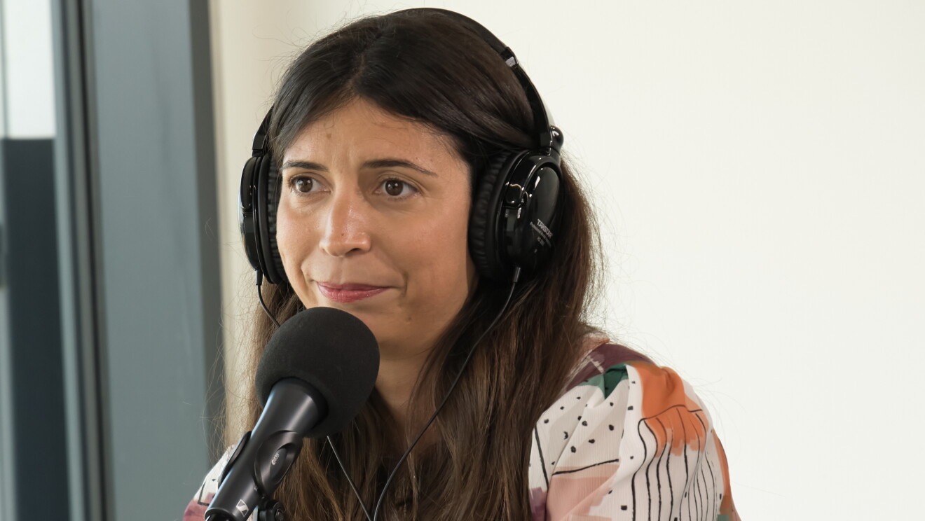 Amanda Machado, CEO de Andrés Machado, sentada en una mesa delante de un micrófono y con unos auriculares puestos. Ella está mirando al infinito. Lleva el pelo largo con pelo castaño. Con una camisa  estampada con puntos y cuadrados dibujados y pinceladas de colors como morado, verde, rosa o naranja.