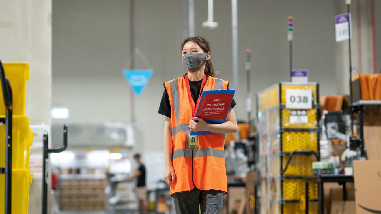 An associate is walking on the floor at Amazon's Fulfillment Center in Singapore with a safety mask on and a file in her hand