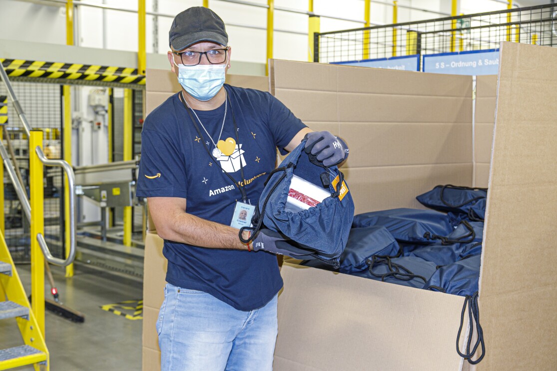 Amazon Helfer vor einem großen Karton mit Rucksäcken.