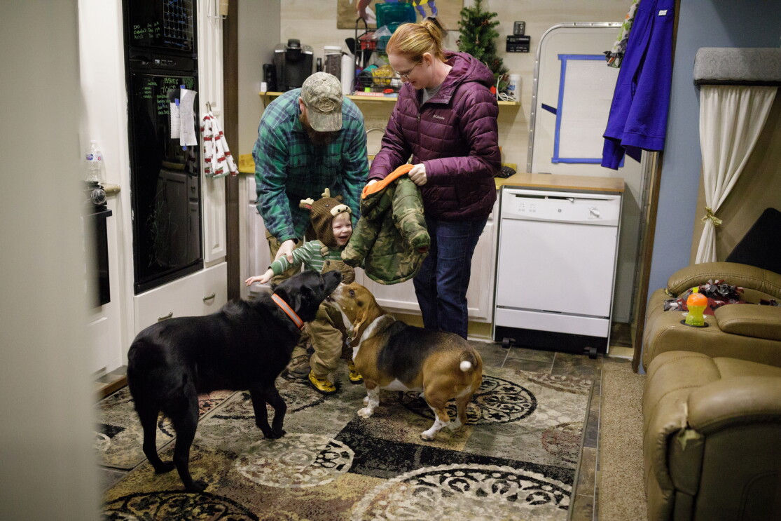 A man, a woman, and a toddler stand inside a home. The woman holds a small jacket. Two dogs are in the foreground.