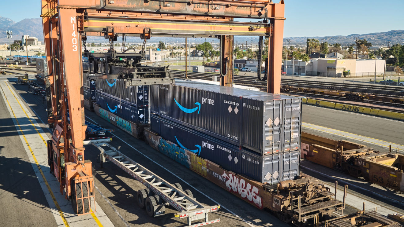 An orange gantry crane loads Amazon Prime containers onto a rail car at a facility.