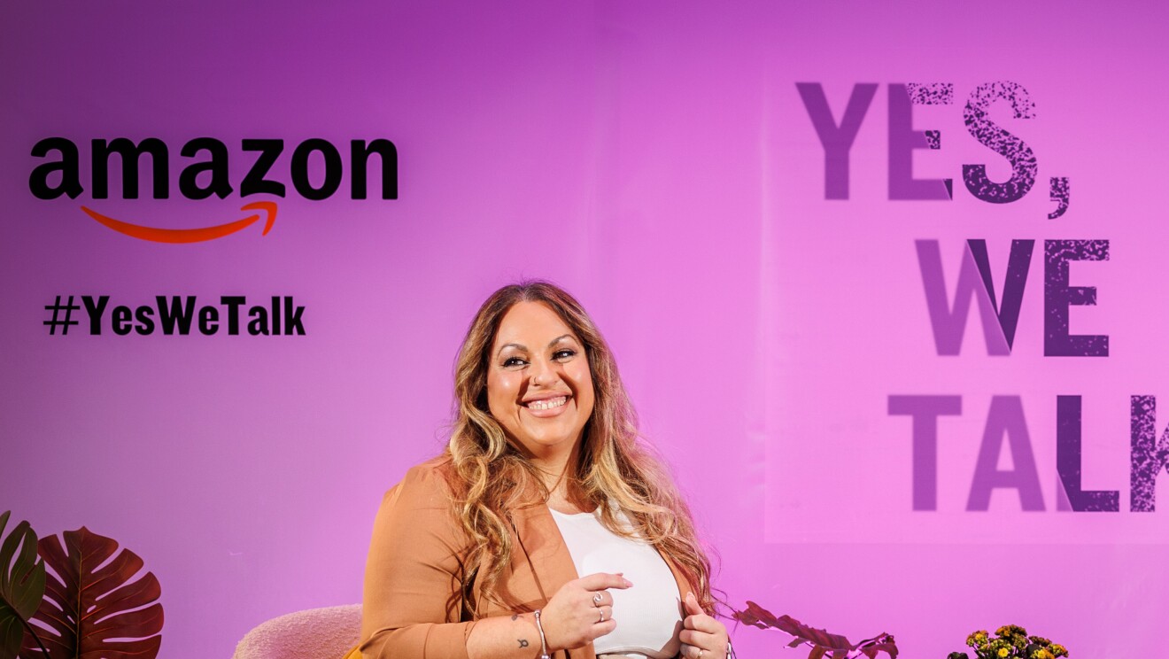 En un escenario creado para la ocasión, de fondo hay un cartel con el logo de Amazon, el Yes, we talk, que es el lema de la campaña y el lema de la sesión Rompiendo el silencio. Todo el fondo es de color rosa. Laura, una trabajadora de Amazon, está hablando agarrando con la mano izquierda unos papeles. Lleva el pelo largo, ondulado, castaño con mechas rubias, Va con tonos beige. Un pantalón de cintura alta, americana marrón clarito y jersey blanco. lleva un brazalete en cada mano y también tatuajes en ambos brazos.