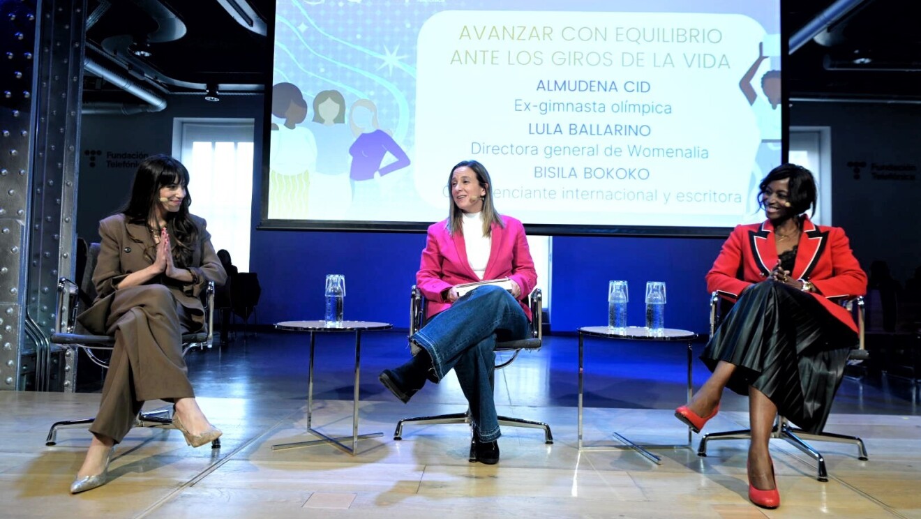 Sentadas en un escenario aparecen Almudena Cid, exgimnasta olímpica, Lula Ballarino, directora general de Womenalia y Bisila Bokoko, emprendedora, escritora y filántropa hispano-estadounidense, entre otras personalidades. Están sentadas en una silla grises y negras. Entre las tres, hay dos mesas con botellas de agua. De fondo una pantalla donde aparecen sus nombres y sus cargos y el nombre del programa Mujeres Que Rompen barreras. Almudena Cid lleva un traje chaqueta de color marrón. Lula va con unos jeans, un jersey de punto blanco y de cuello alto y una americana rosa. Bisila va con una falda negra imitación piel, un blazer de color negro y una americana roja, con motivos blancos y negros. Bisila es afrodescendiente.
