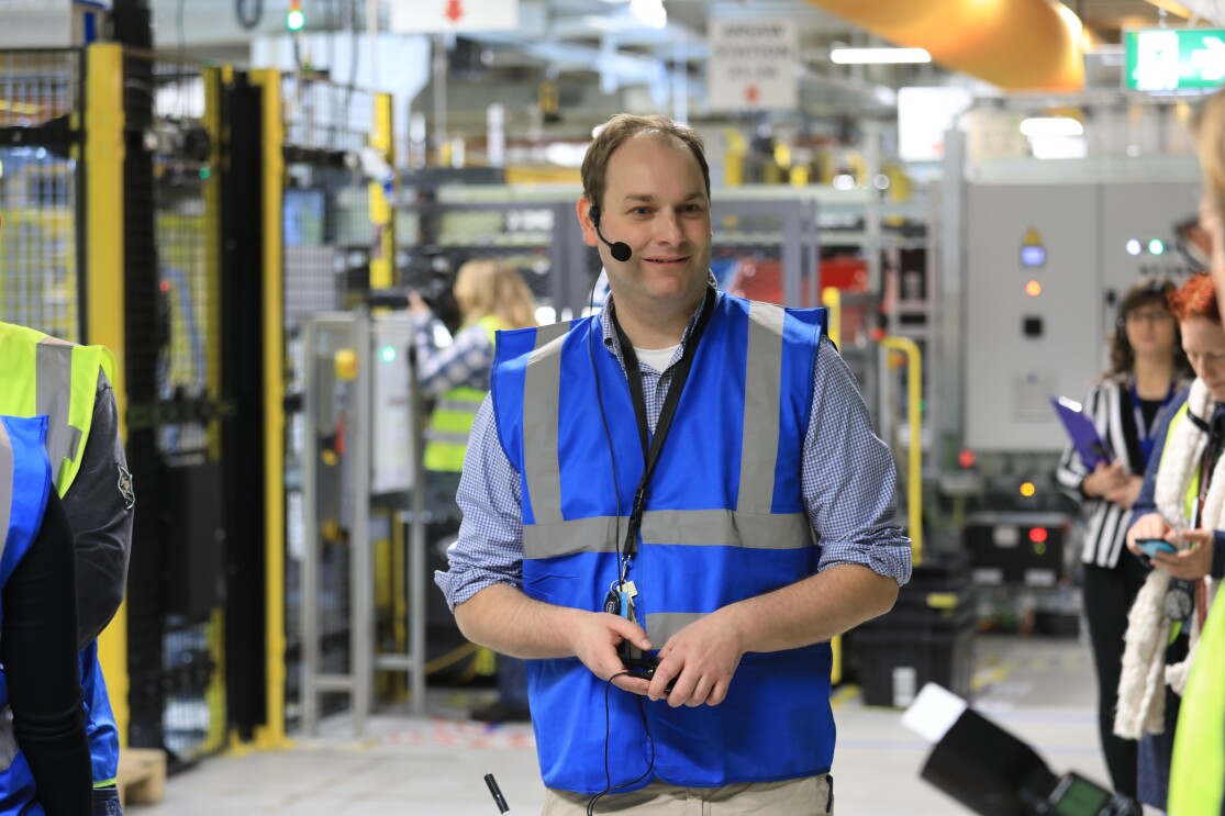 Ein Mann mit blauer Sicherheitsweste steht in einer Logistikhalle, hinter ihm sieht man Besucher