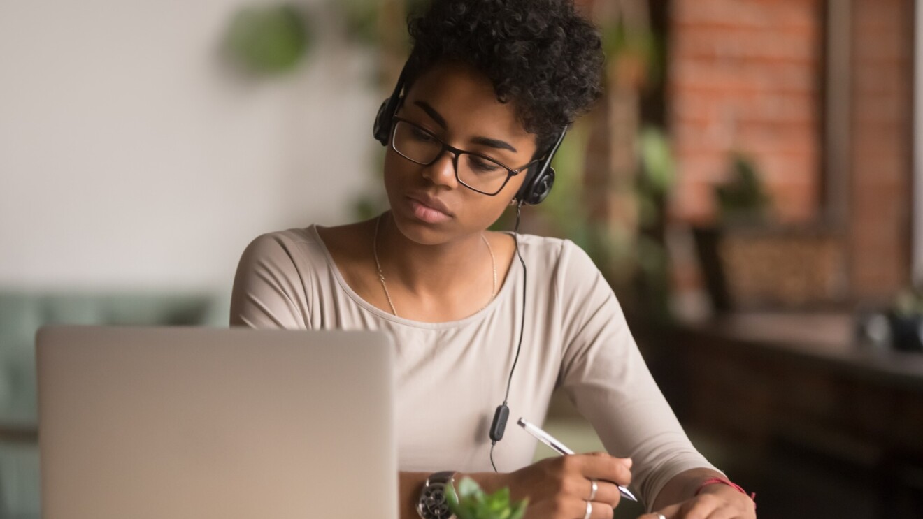 Woman watches her laptop while taking notes. She is wearing glasses and a headset.