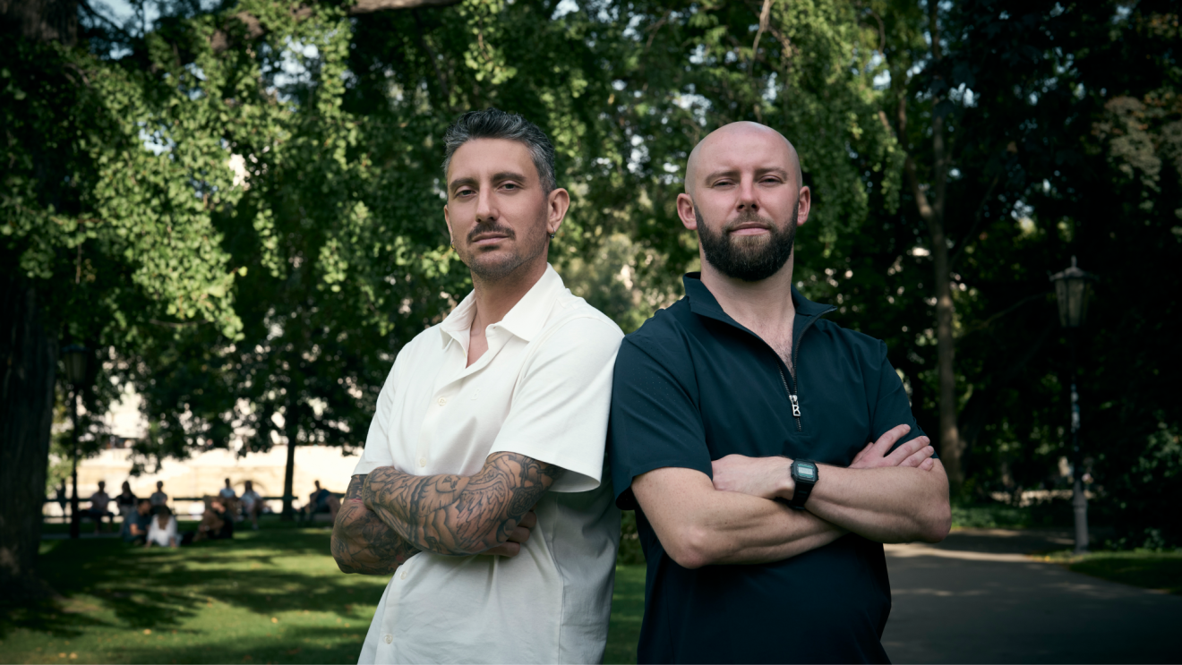 Two men folding their arms while standing in a park and looking into camera